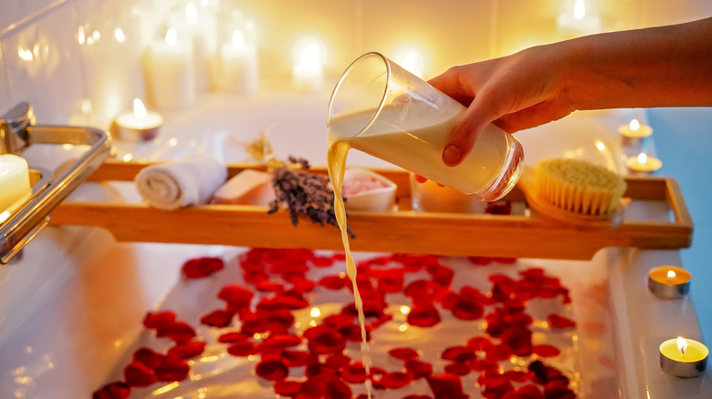 Woman's hand pouring glass of coconut milk into bath water with rose petals