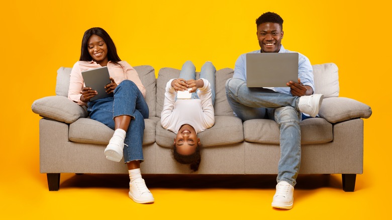 woman, man, and young girl sitting on couch smiling at their electronics