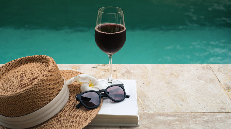 book next to a pool with a hat, sunglasses, and glass of wine