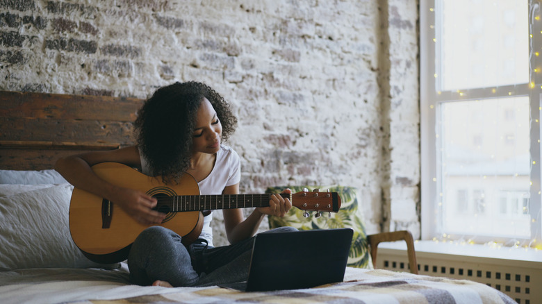 Woman playing guitar in bed looking at her laptop