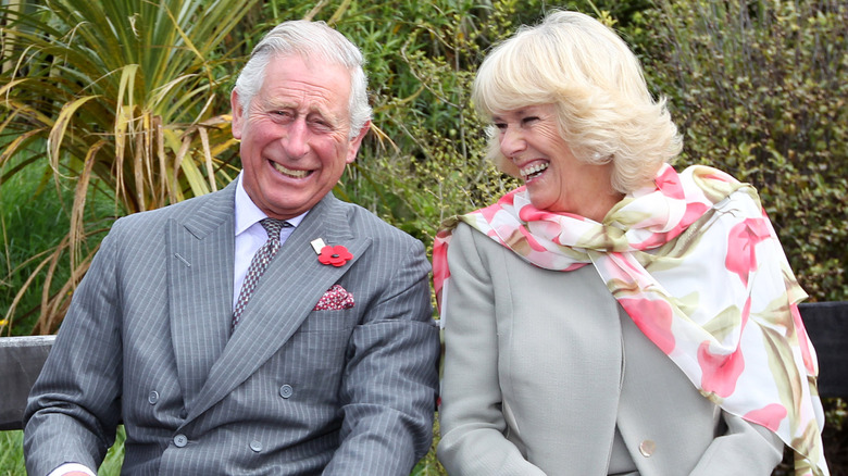 King Charles and Queen Camilla sitting next to each other on bench and laughing