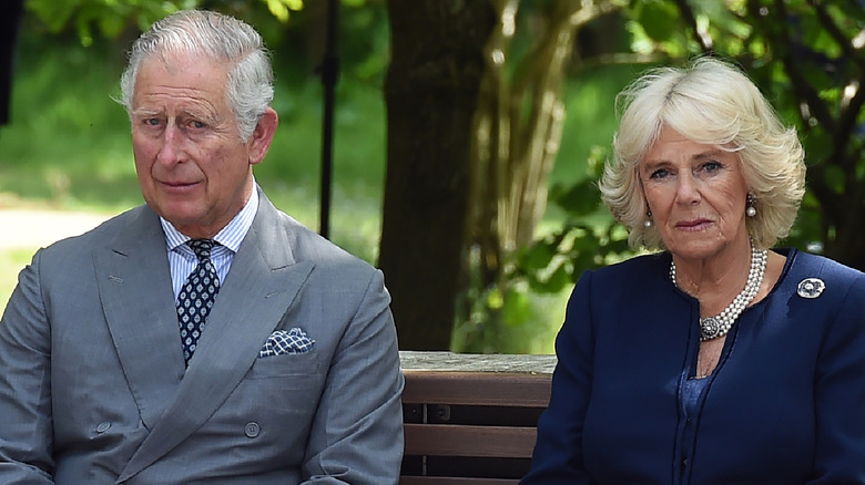 King Charles and Queen Camilla looking serious sitting on a bench surrounded by trees