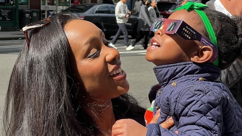 Abby Phillip smiling while holding daughter Naomi, who wears eclipse glasses