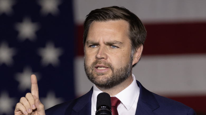 JD Vance pointing up while speaking with an American flag behind him