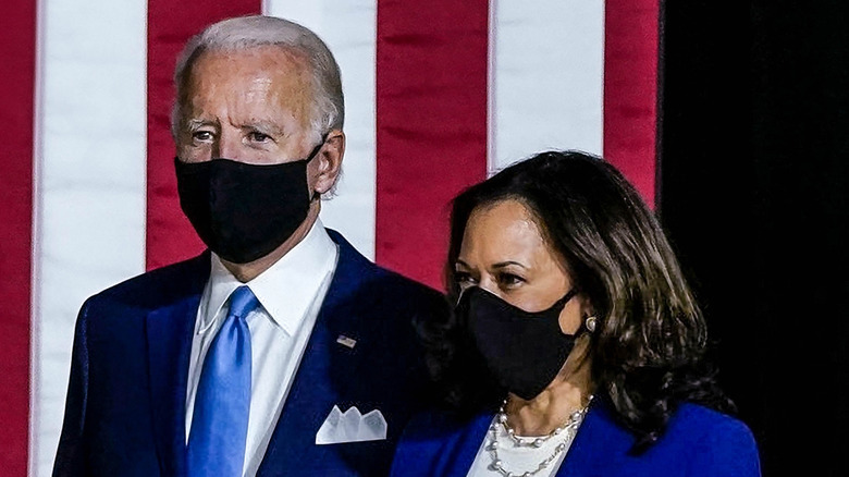 President Joe Biden and Vice President Kamala Harris standing together and looking to the left. Both wear blue suits and black face masks against a backdrop of the American Flag