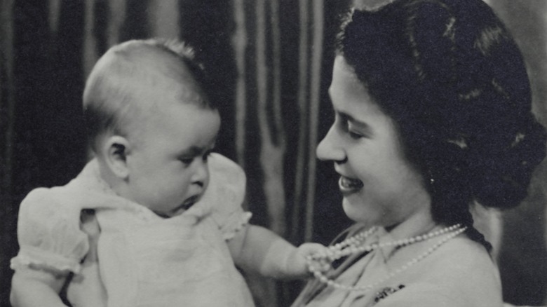 Queen Elizabeth holding Prince Charles as a baby who's playing with her pearls