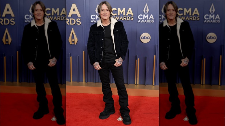 Keith Urban on the CMA Awards red carpet wearing black jeans, black T-shirt, clunky shoes, and a fuzzy-lined jacket.