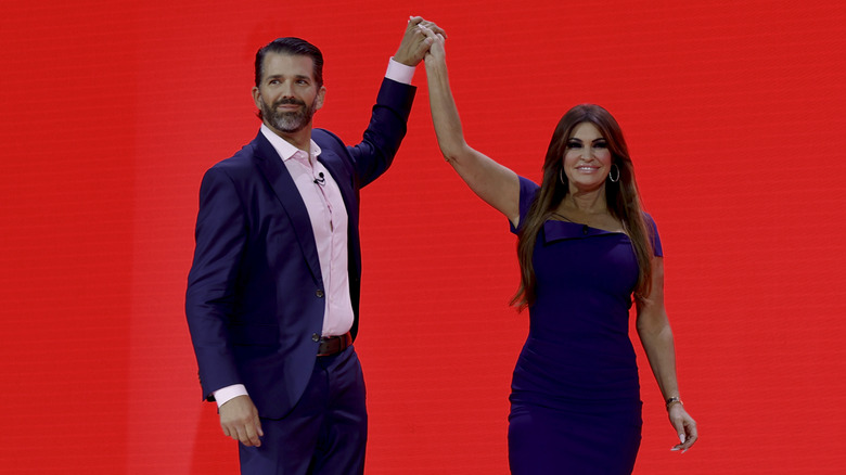 Donald Trump Jr. and Kimberly Guilfoyle hold hands while standing against a red background