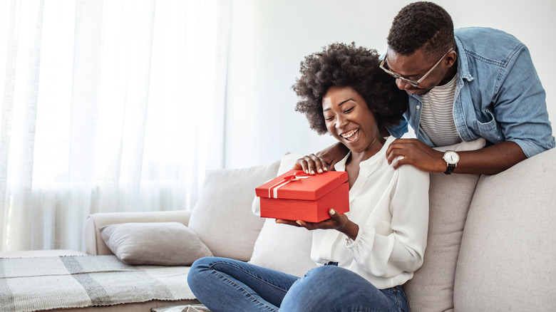 Man gives woman gift while she sits on sofa