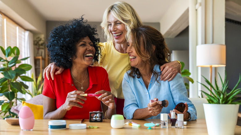 Three women laughing and embracing while doing makeup