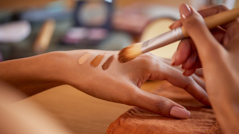 Woman holding makeup brush in one hand while different shades of foundation makeup are on her other hand