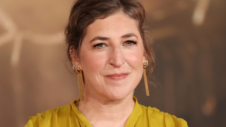 Mayim Bialik smiling in a yellow dress at a press event