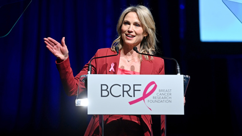 Amy Robach smiles and waves behind podium and a sign that reads "Breast Cancer Research Foundation" at an event