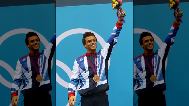 Tom Daley smiling while holding up flowers and wearing a bronze medal