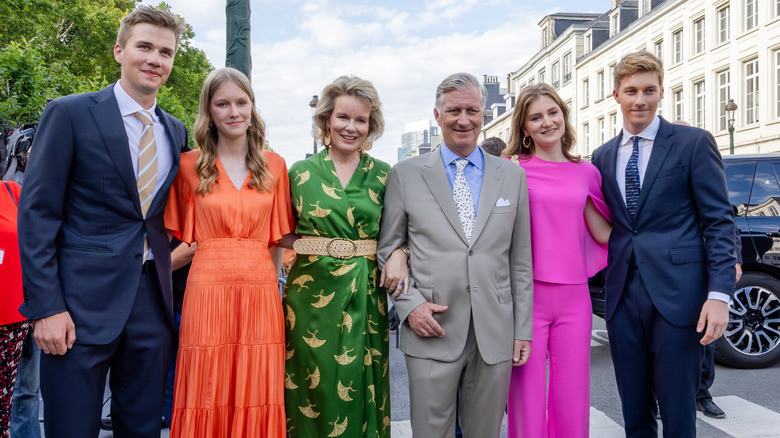 The royal family of Belgium stand together on a sidewalk posing with arms linked.