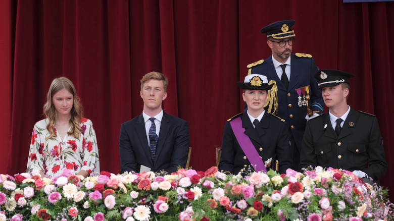 Prince Gabriel of Belgium sits between his siblings in front of a red curtain.