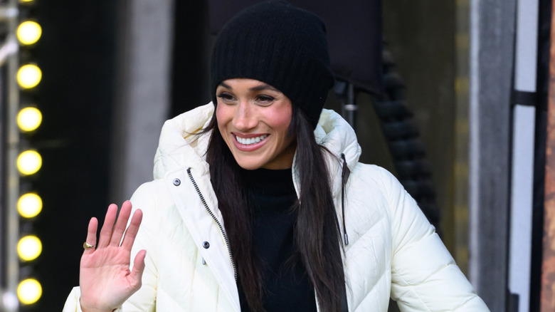 Meghan Markle smiles, wearing a white puffy coat and a black beanie