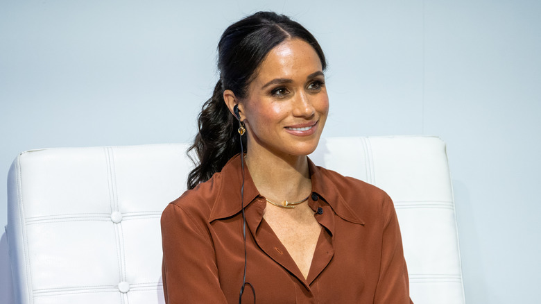 Meghan Markle smiling onstage in a brown blouse