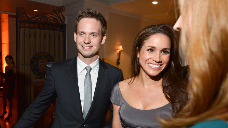 A smiling Meghan Markle walking with Patrick J. Adams smiling in a suit behind her