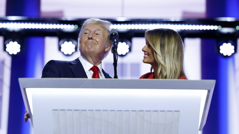 Melania Trump smiling at Donald Trump behind podium