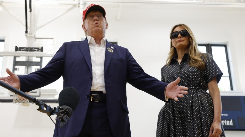 Donald Trump speaking, while Melania Trump stands nearby in sunglasses