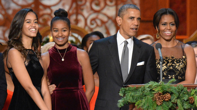 Malia, Sasha, and Michelle Obama smiling while Barack Obama speaks