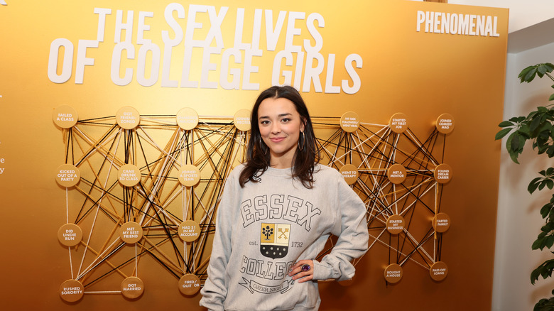 Midori Francis smiling in front of promo for The Sex Lives of College Girls