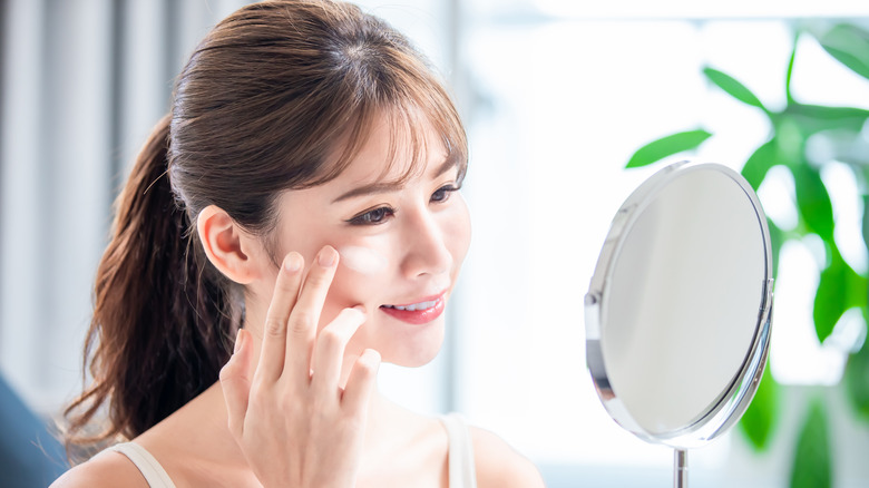 Brunette woman applying sunscreen to her face while looking in the mirror.