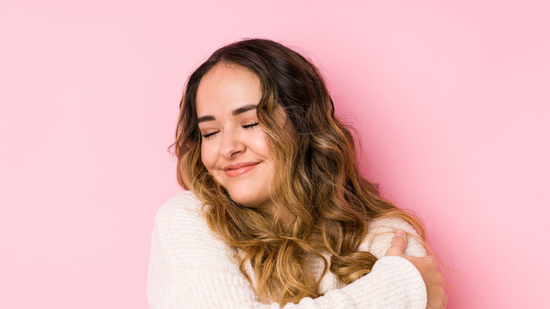 person hugging herself in front of pink wall