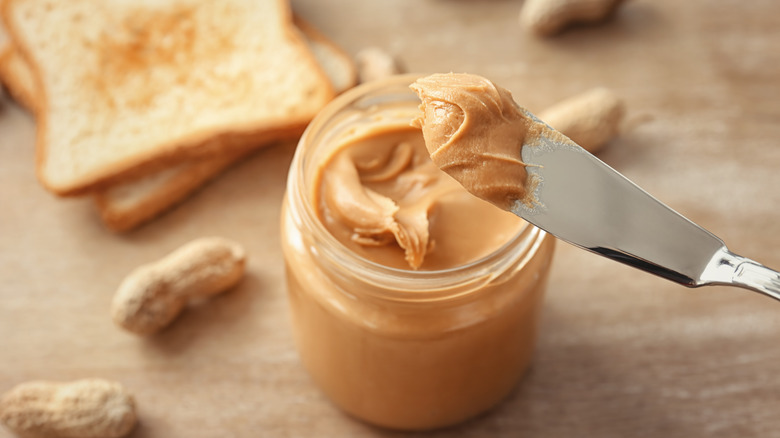 A dollop of peanut butter on a knife with a jar of peanut butter below it