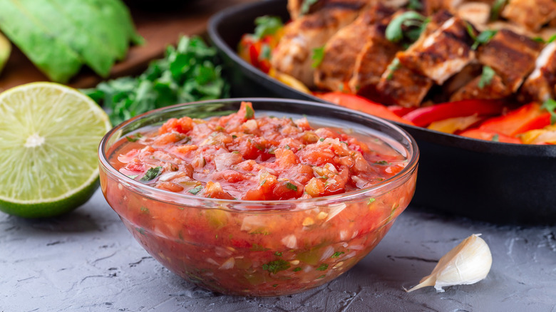 A bowl of salsa surrounded by other dishes, a lime, avocados, and garlic