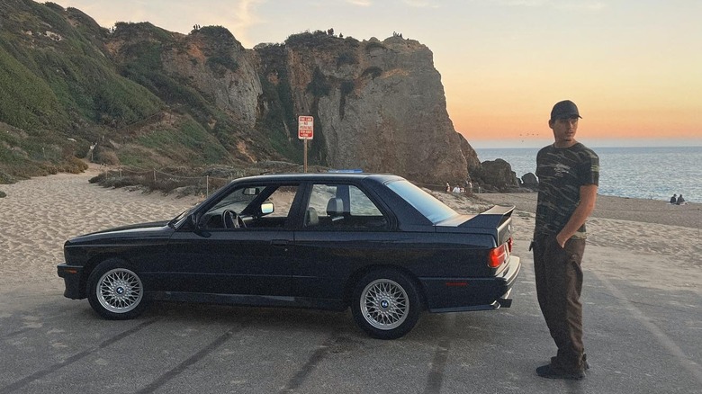 Paris Brosnan stands outside near the beach next to his BMW.