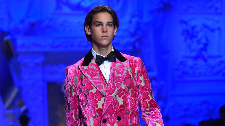 Paris Brosnan walks the runway during a Dolce and Gabbana show wearing a pink pattern suit jacket.