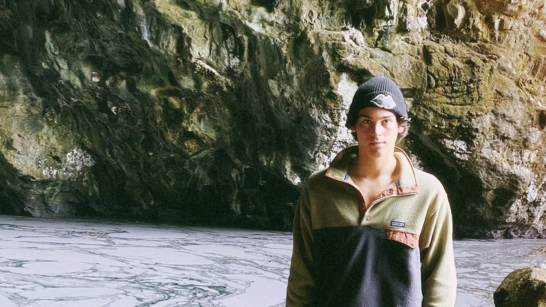 Paris Brosnan faces the camera wearing a beanie outside a cave.