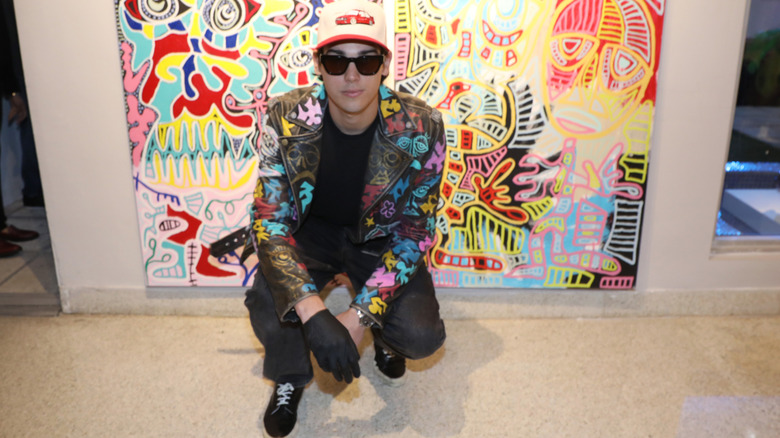 Paris Brosnan squats in front of a colorful canvas of his artwork at a gallery show.