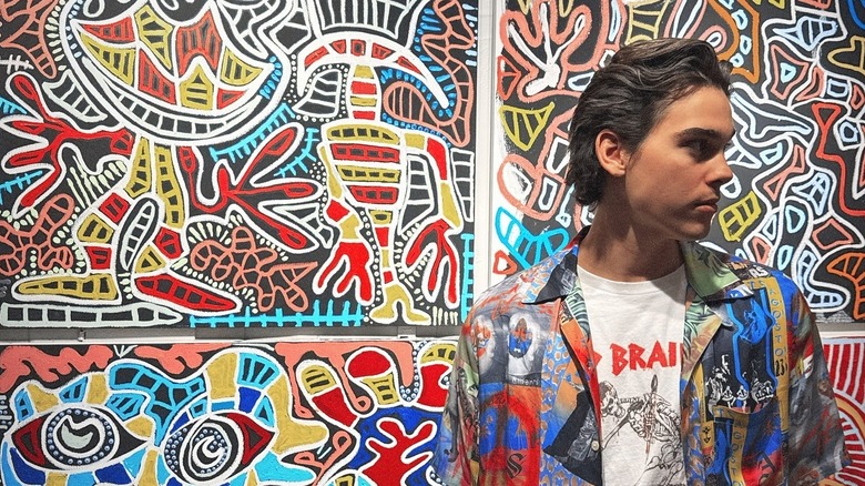 Paris Brosnan turns his head to the side standing in front of his colorful paintings in a bright pattern shirt.