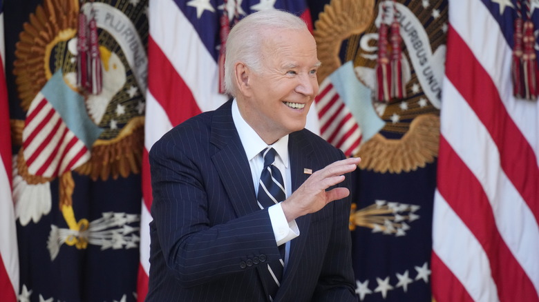 Joe Biden smiling and waving in front of several flags