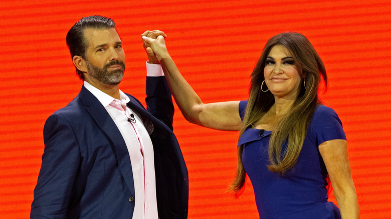 Kimberly Guilfoyle and Donald Trump Jr. holding hands in front of a red background