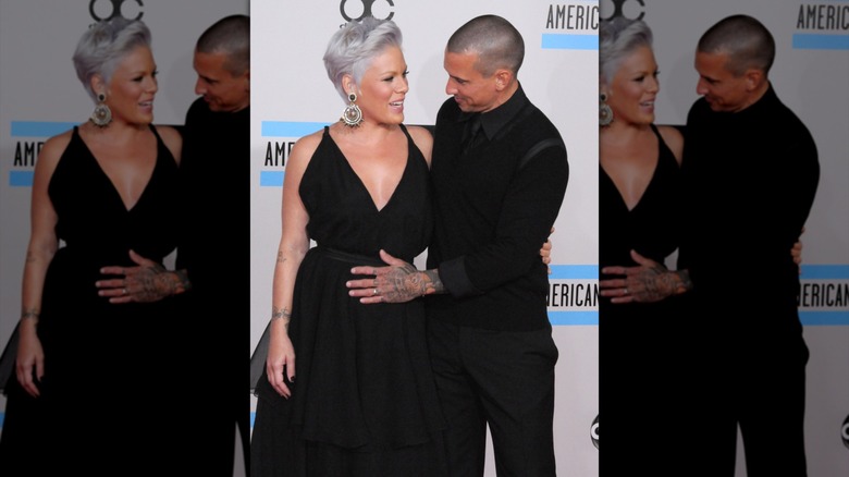 Pregnant Pink with short gray hair in black dress and Carey Hart in black with his hand on her belly