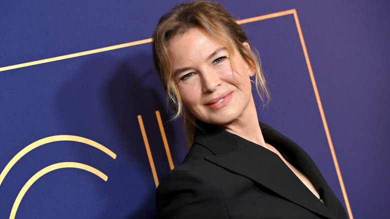 Renée Zellweger wearing a black blazer, her head tilted as she looks at the camer and smiles.