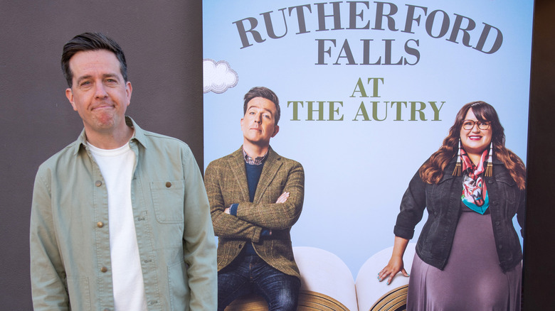 Ed Helms posing next to a "Rutherford Falls" poster