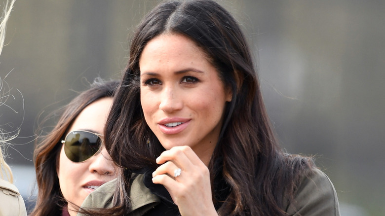 Meghan Markle smiling with her hand raised.