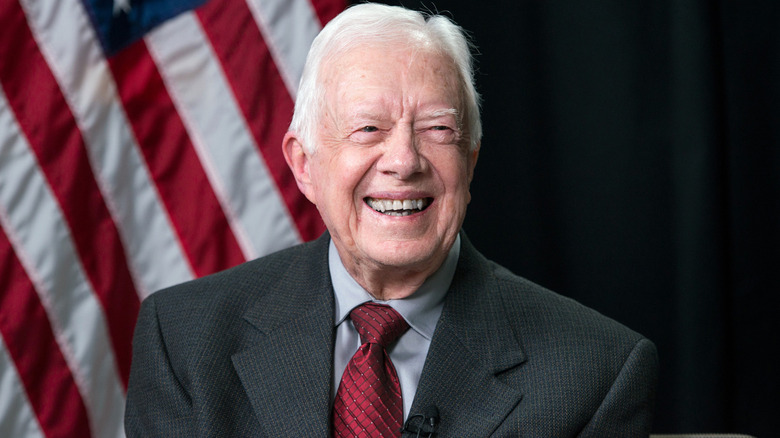 Former US President Jimmy Carter with a big smile, looking off the side, an American flag in the background.