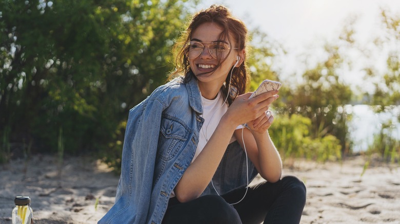 Young brunette woman sitting on beach, smiling and listening to earphones