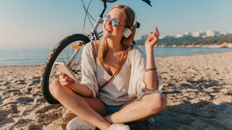 White woman laughing, sitting on beach, and listening to headphones