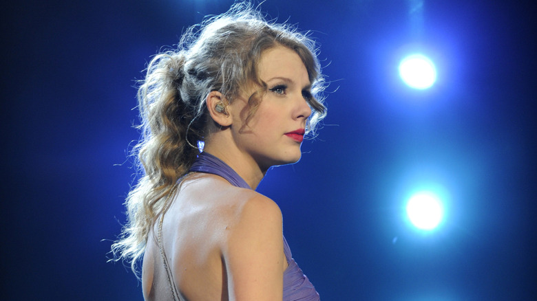 Taylor Swift on stage during Speak Now tour in purple dress, red lipstick, and wavy ponytail