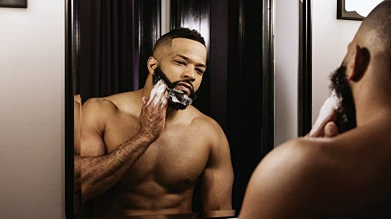 Shirtless man grooming his beard in the mirror