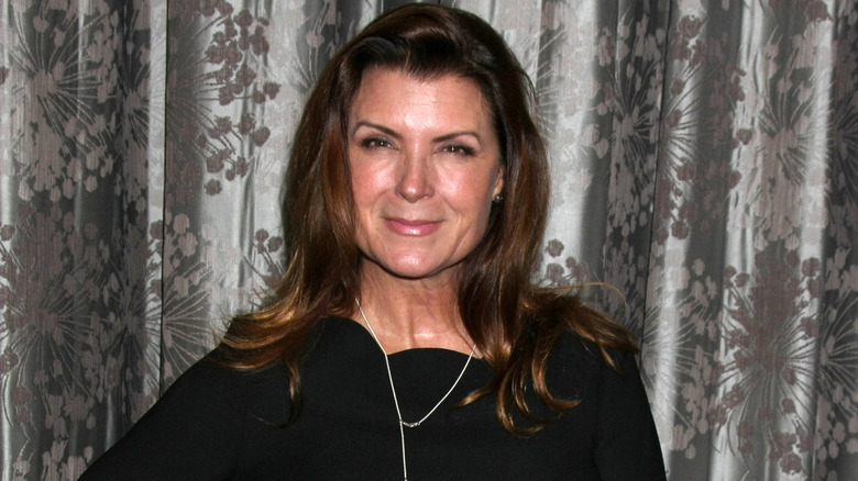 Actress Kimberlin Brown poses in front of a gray background while wearing a black dress.