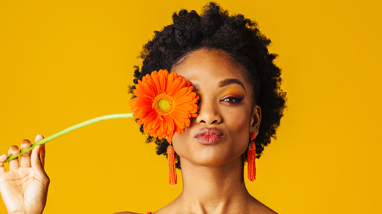 Woman with orange eyeshadow and orange lipstick covering one eye with flower