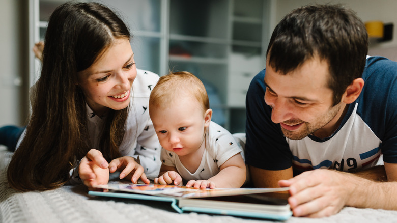 mother, father, and baby lying in bed and looking at book